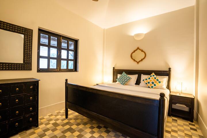 Relax in this inviting bedroom at our Goa villa, offering a comfortable bed, stylish dark wood furniture, and charming details for a peaceful stay.