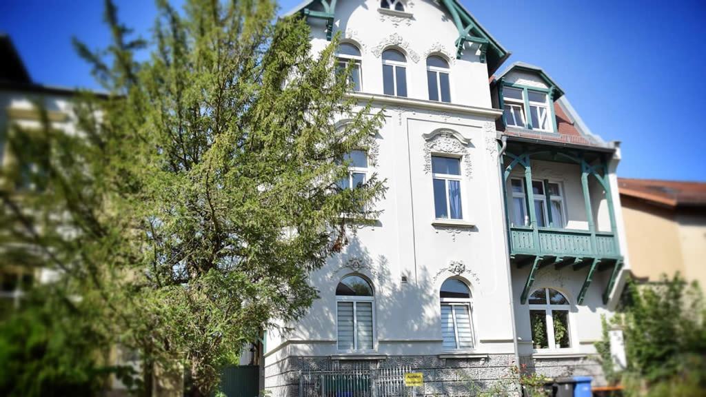A historic villa showcases an elegant facade with decorative architectural details. Large windows allow natural light to fill the interior. A blend of green accents can be seen in the balcony's trim and the lush greenery surrounding the building, providing a welcoming exterior.