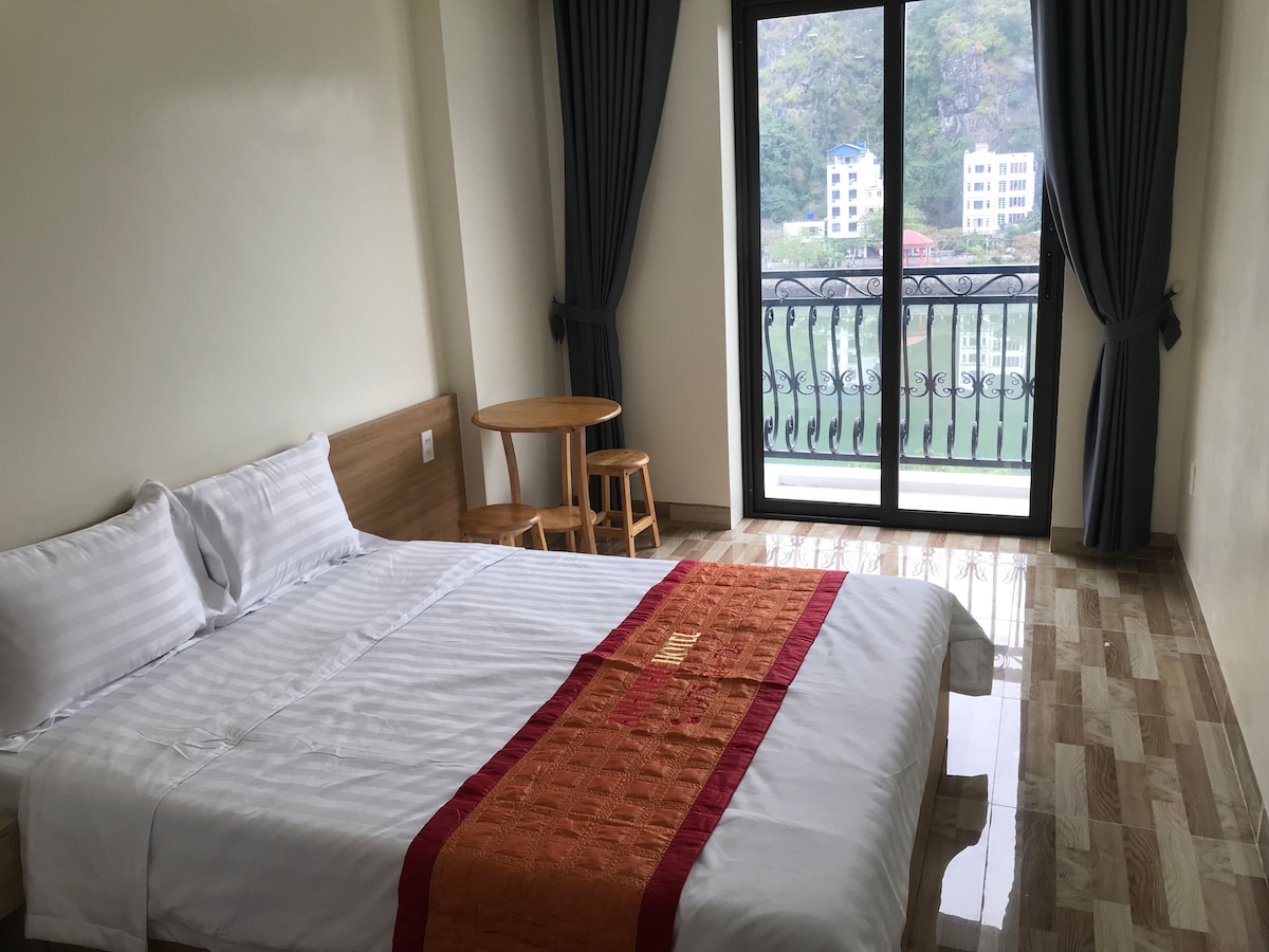 A bright room features a spacious bed with fresh linens and a decorative red runner. A small wooden table with two stools is positioned by the floor-to-ceiling balcony doors, which provide a view of the surroundings. Light-colored tiles enhance the airy feel of the space.