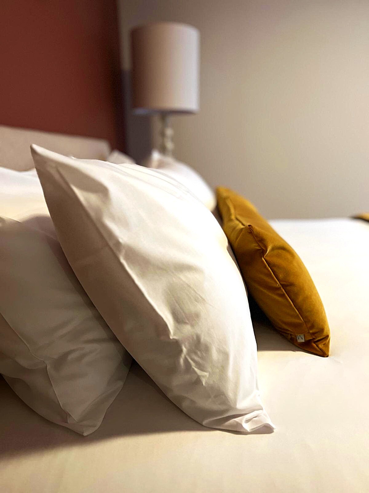 A spacious bed is complemented by several neatly arranged pillows, including white and a vibrant mustard-colored pillow. Soft lighting from a nearby lamp softly illuminates the scene, creating a serene and inviting ambiance.