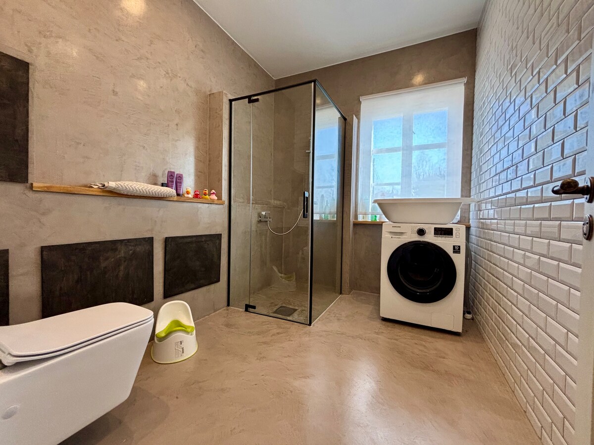 A modern bathroom features a walk-in shower enclosed by glass panels. A freestanding bathtub is situated next to a window, allowing natural light to enter. The room displays a combination of smooth and textured wall surfaces, along with practical amenities, including a washing machine.