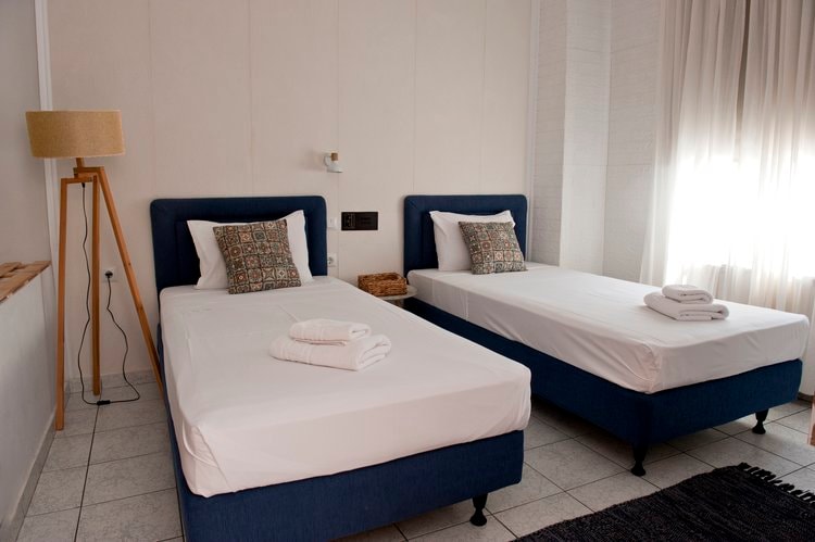 A well-organized guest room features two spacious beds, each adorned with decorative pillows. Soft towels are neatly placed at the foot of the beds. Natural light filters through sheer curtains, illuminating the light-colored walls and floor tiles.