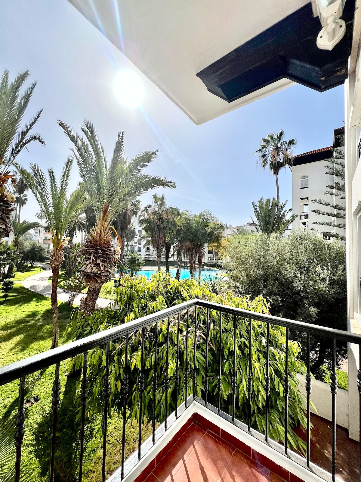 A view from a balcony showcases lush palm trees and vibrant greenery. A glimmering pool is visible amid the foliage, with sunlight reflecting off its surface. The composition highlights the serene outdoor surroundings, creating a tranquil sense of space.