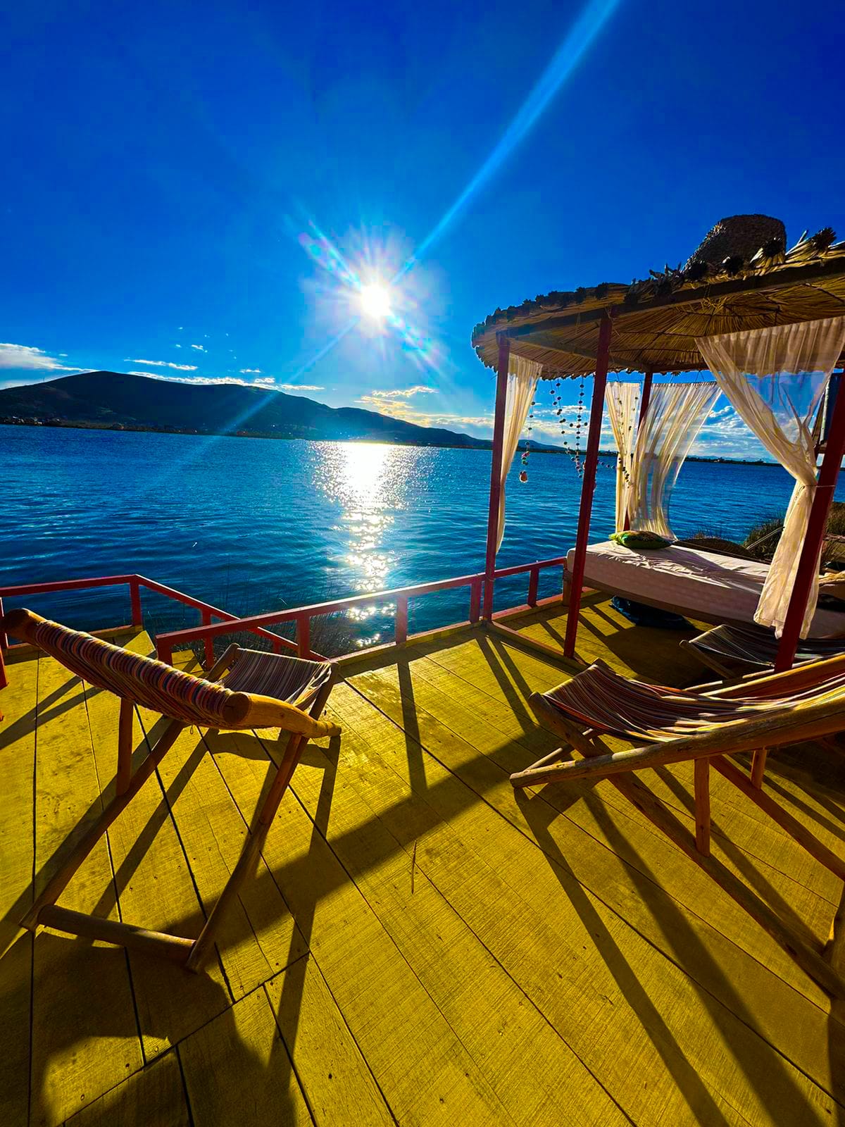 A tranquil outdoor seating area features two wooden loungers positioned on a sunlit deck overlooking the water. A shaded daybed is draped with sheer fabric, enhancing the serene atmosphere. Sunlight reflects off the lake, and hills are visible in the background.
