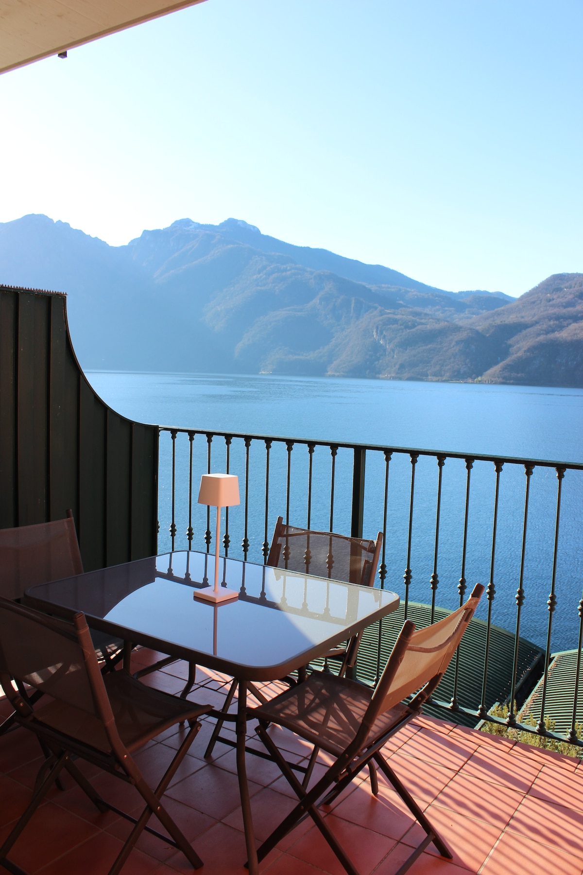 A balcony featuring a glass table surrounded by four chairs offers a panoramic view of the shimmering lake and mountains in the background. A small lamp provides soft lighting, enhancing the serene outdoor setting, perfect for relaxation or dining.
