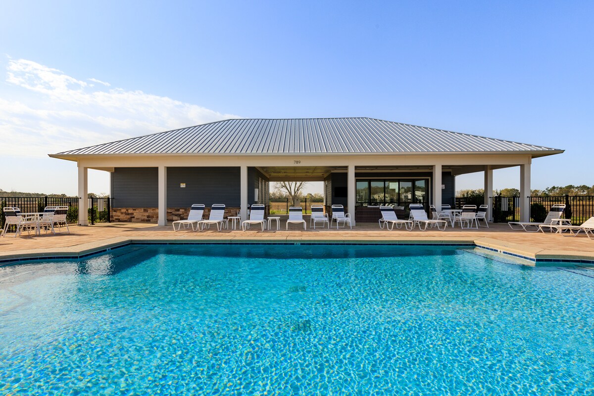 A pool area features a sparkling blue pool surrounded by white lounge chairs. A spacious clubhouse with large windows overlooks the pool, providing ample seating. The clear sky is visible above, and the landscape includes greenery in the background, creating a relaxing environment.