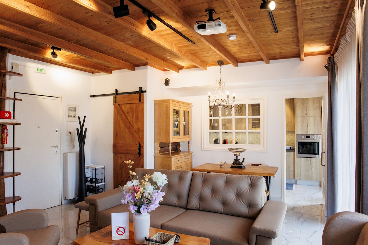 A bright living room features wooden beams on the ceiling and a spacious sofa with a light-colored upholstery. A dining area with a wooden table is visible in the background, while a projector screen is mounted on the wall, enhancing the room's entertainment options.