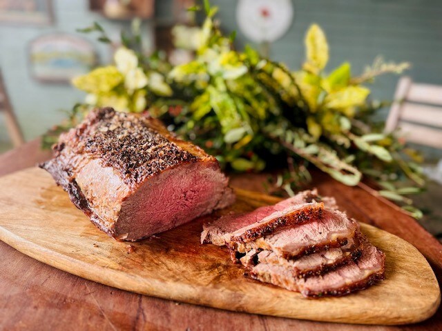 A beautifully presented roast sits on a wooden cutting board. The meat displays a perfectly cooked pink interior, with slices arranged neatly next to it. Fresh green foliage in the background adds a natural touch to the scene.