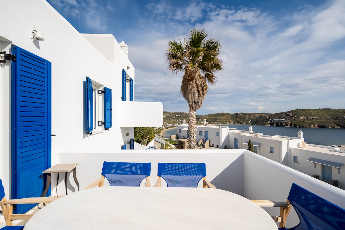 A spacious terrace is furnished with a round table and four blue chairs, offering a relaxing place to enjoy the outdoor ambiance. White walls and blue shutters frame the view of the Aegean Sea and surrounding landscape, accentuated by a tall palm tree swaying gently.