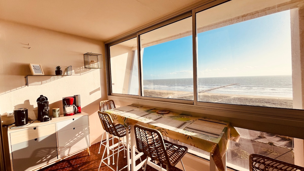 A bright dining area features a long table set against expansive windows, providing a view of the ocean. High-backed chairs surround the table, while a well-equipped kitchen corner is visible with various appliances arranged neatly on a sideboard.