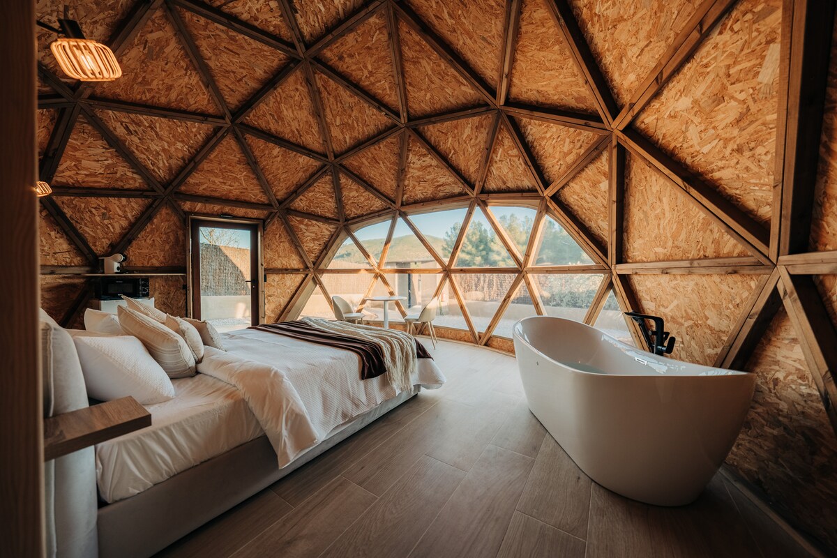 A spacious dome interior features large triangular windows allowing natural light to brighten the room. A comfortable bed is presented alongside a freestanding bathtub. A small dining area is visible, enhancing the cozy layout with wooden accents throughout.