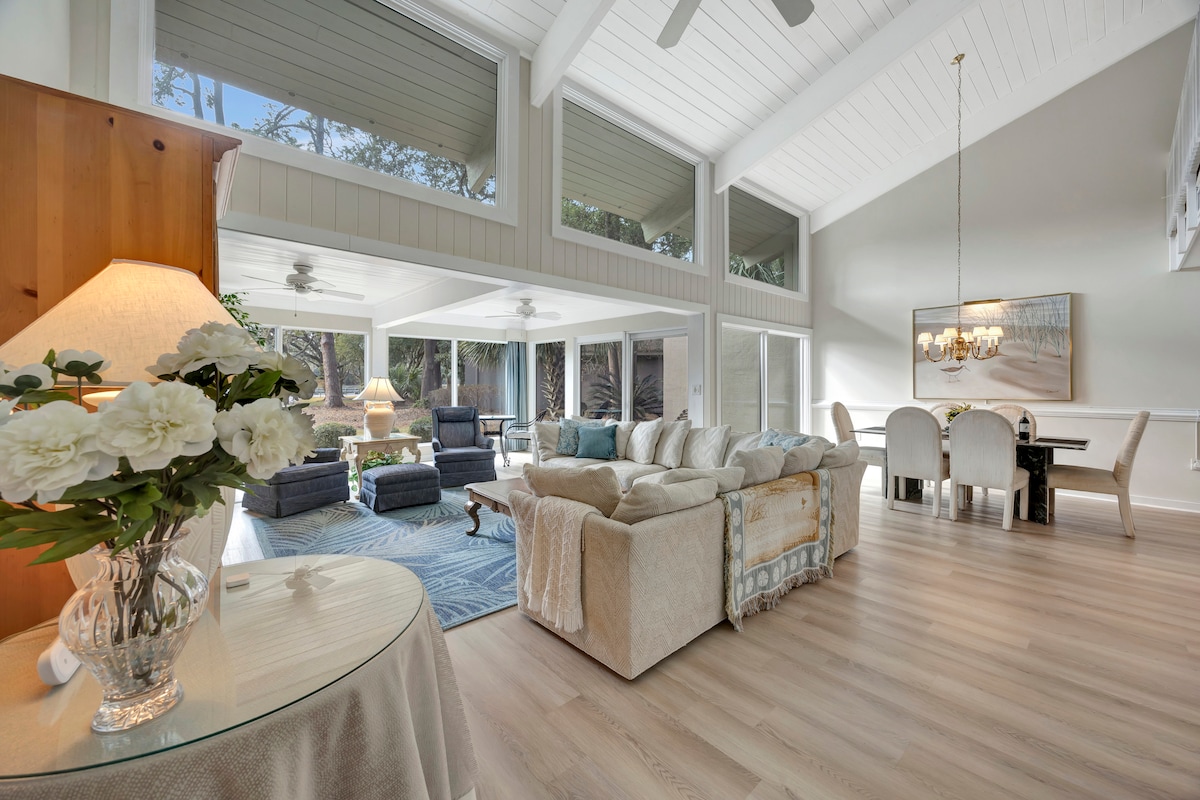 An airy living area features tall ceilings and large windows, allowing natural light to fill the space. Comfortable seating arrangements are present alongside a stylish coffee table. A dining area with a chandelier can be seen in the background, completing the inviting layout.