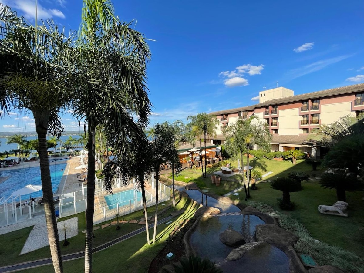 A view of the resort grounds showcases a well-maintained green area with palm trees and landscaped paths. The pool area is visible, accompanied by lounge seating. The lake can be seen in the background under a bright blue sky, contributing to a serene outdoor atmosphere.