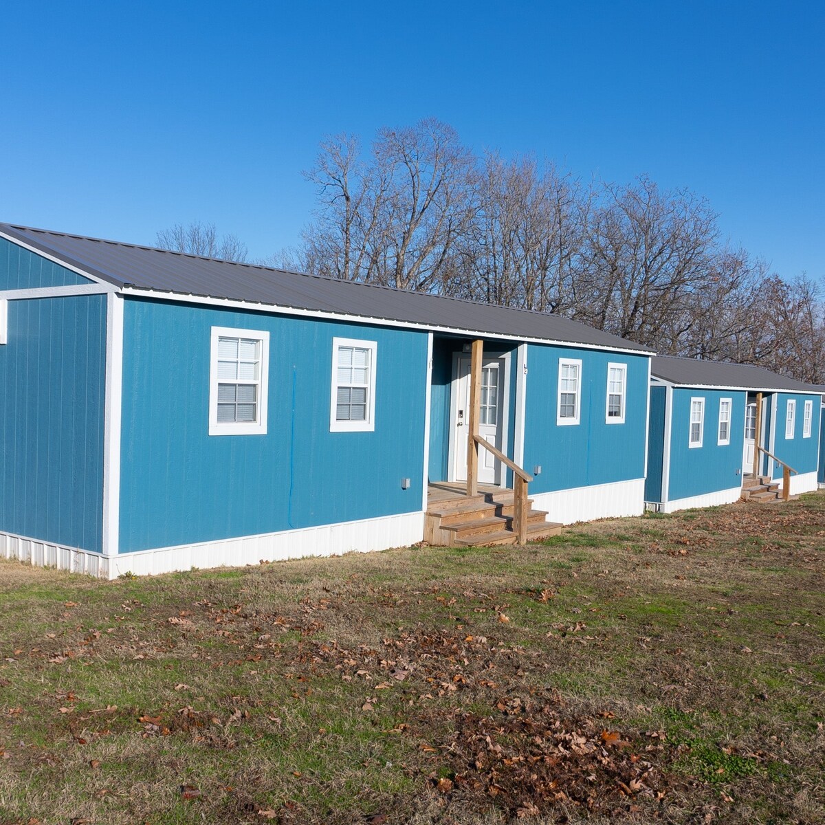 Two duplex-style cabins are visible, each featuring a blue exterior and a simple porch with wooden steps. Large windows are positioned symmetrically along the facade, allowing natural light to fill the interior spaces. A grassy area with fallen leaves surrounds the cabins.