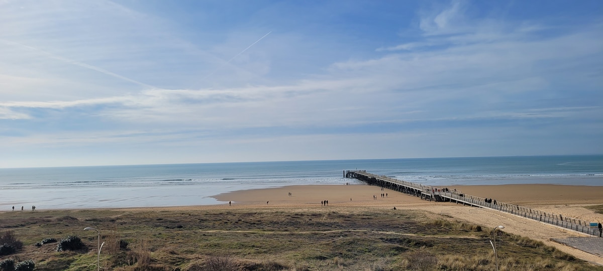 A wide view captures a sandy beach leading to a wooden pier extending into the open sea. Gentle waves lap at the shore, while distant figures stroll along the beach. Soft clouds drift across a blue sky, framing a serene coastal landscape.