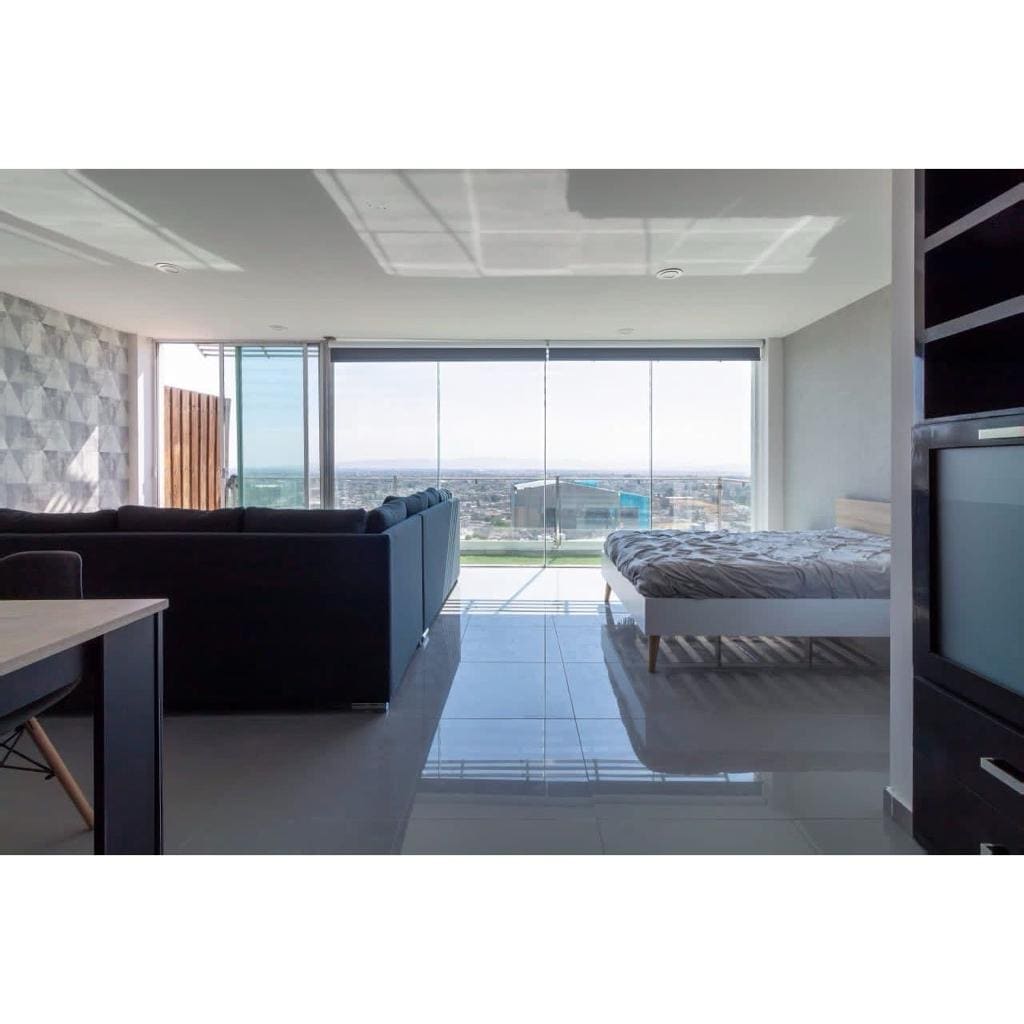 A modern living space is captured, featuring an arrangement of sleek furniture. A spacious bed sits in the background, while a large glass wall provides an expansive view of the city. Natural light enhances the room, reflecting off the polished floor tiles.
