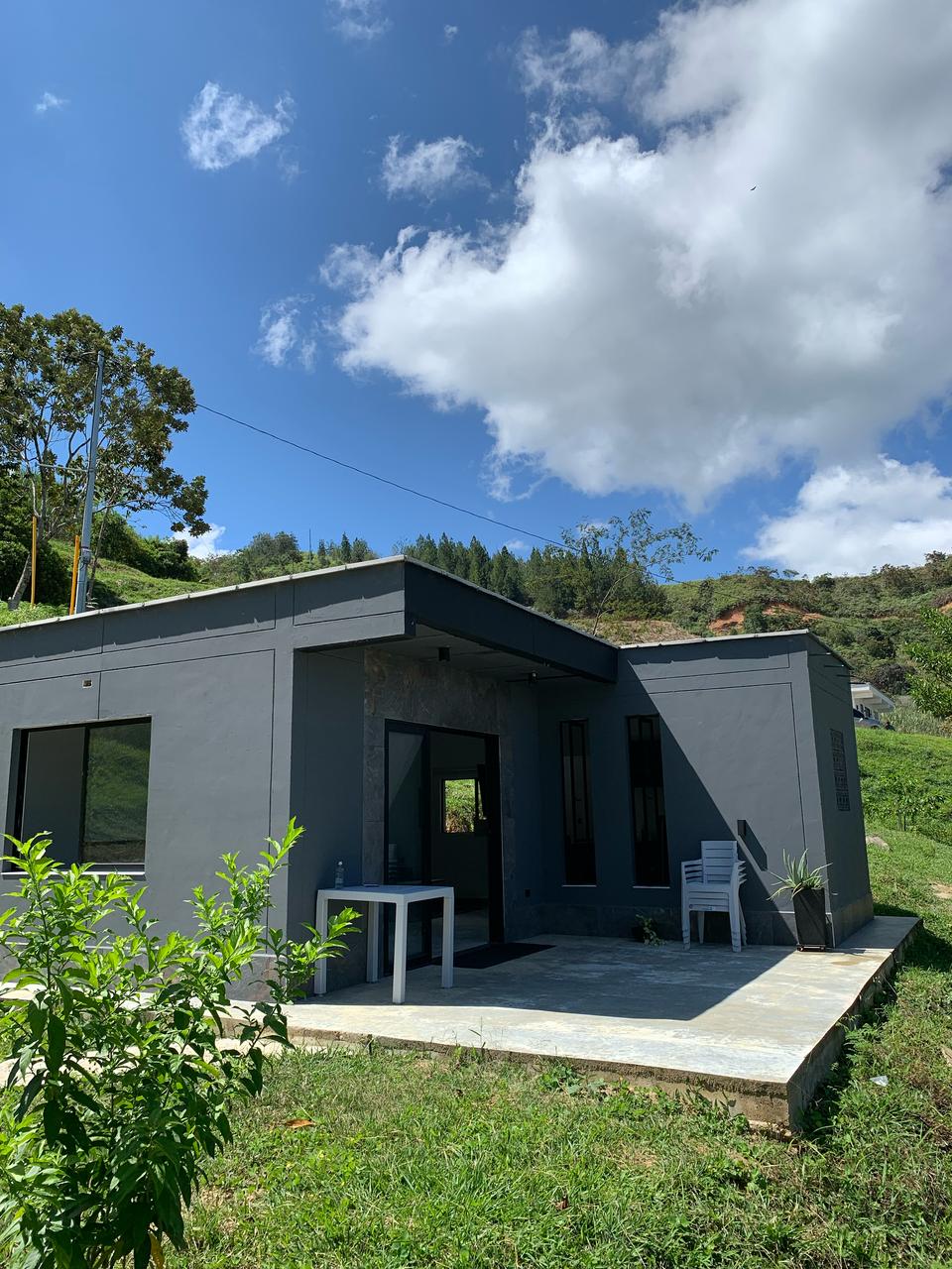 A modern cabin is set amidst lush greenery, featuring a minimalist gray exterior. A small table and a white chair are visible on the stone patio, while large windows provide natural light and views of the surrounding landscape.