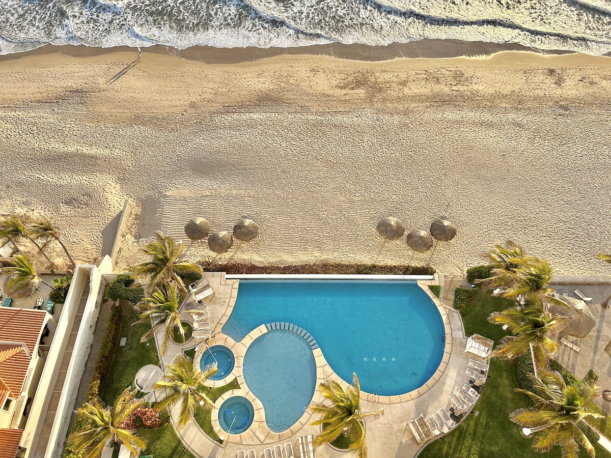 An aerial view captures a spacious pool surrounded by palm trees, with a beach extending into the distance. Lounge chairs are arranged near the pool, providing a serene setting by the waves. Umbrellas are positioned on the sandy beach, enhancing the coastal atmosphere.