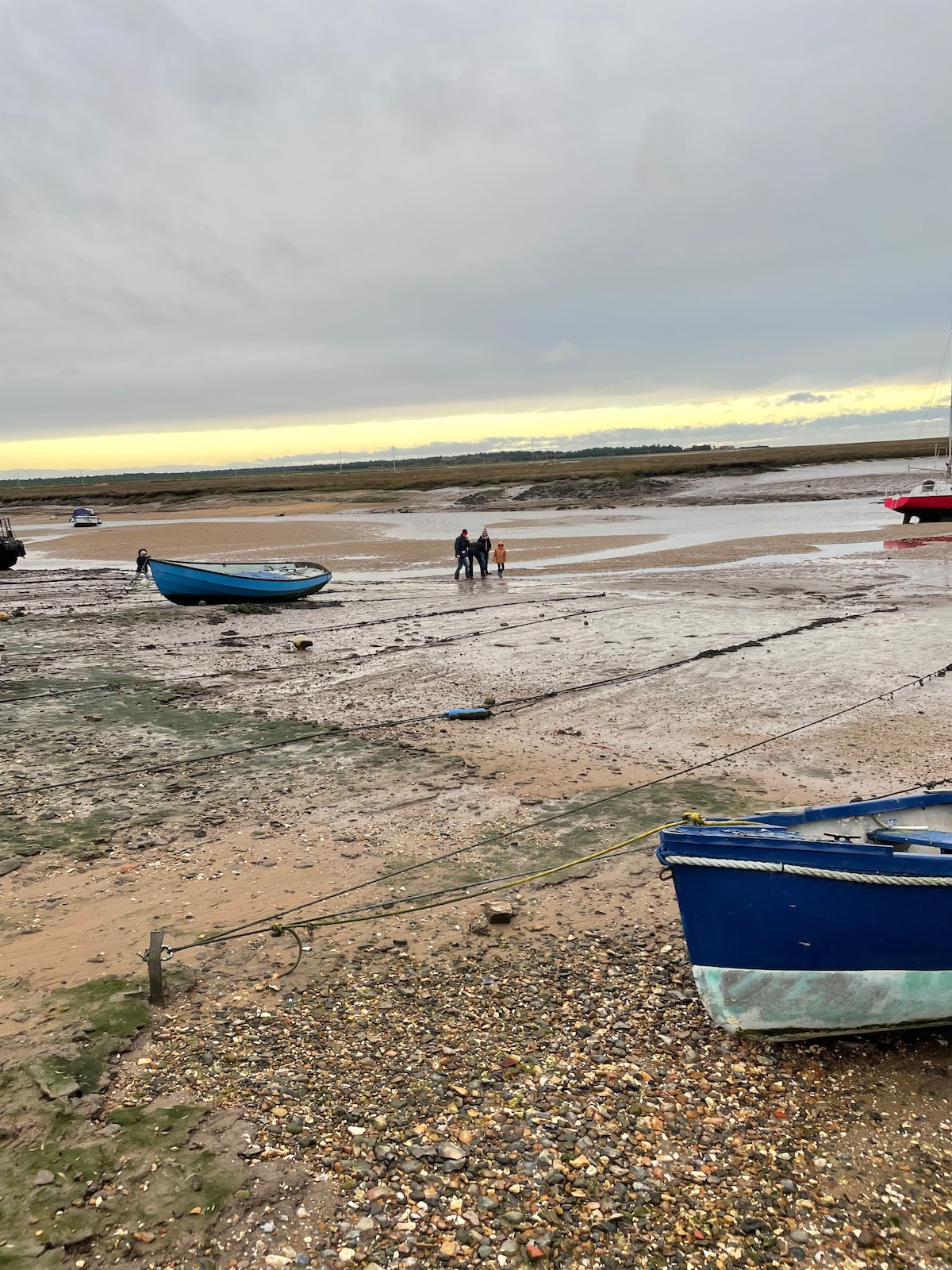 Two individuals are seen walking along a sandy shore during low tide, with boats resting on the ground nearby. The beach features a mix of wet and dry sand, while the horizon is painted with soft hues of orange and grey, suggesting a cloudy sunset.