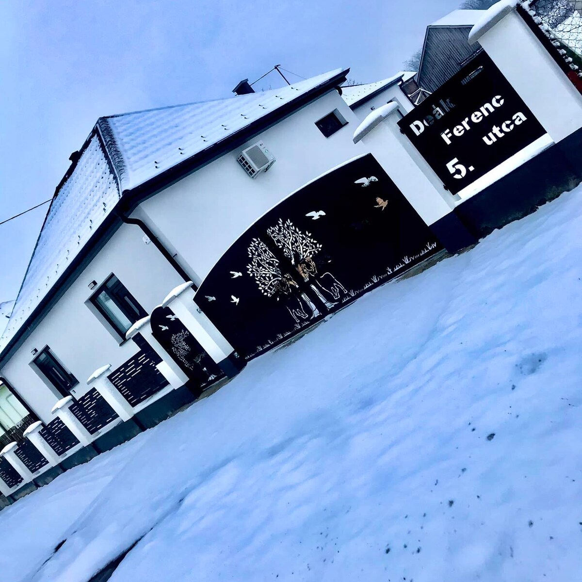 The exterior of the guesthouse is shown, featuring a white facade and a pitched roof blanketed in snow. Artistic tree murals and decorative elements add character to the front wall. Snow covers the ground, creating a serene winter landscape.