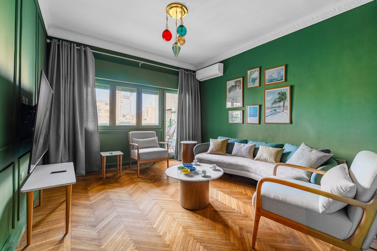 A stylish living area features green walls that create a calming backdrop. Comfortable seating is arranged around a circular coffee table, with natural light filtering through multiple windows. Artwork adorns the walls, adding visual interest to the serene atmosphere.