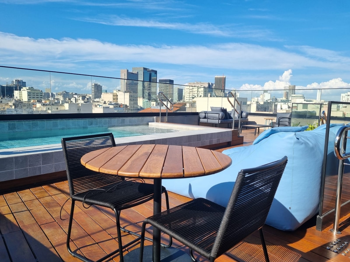 An outdoor rooftop area is highlighted, featuring a wooden table accompanied by four black chairs. A blue bean bag is positioned nearby, with a swimming pool visible in the background. The city skyline stretches across the horizon under a clear blue sky.