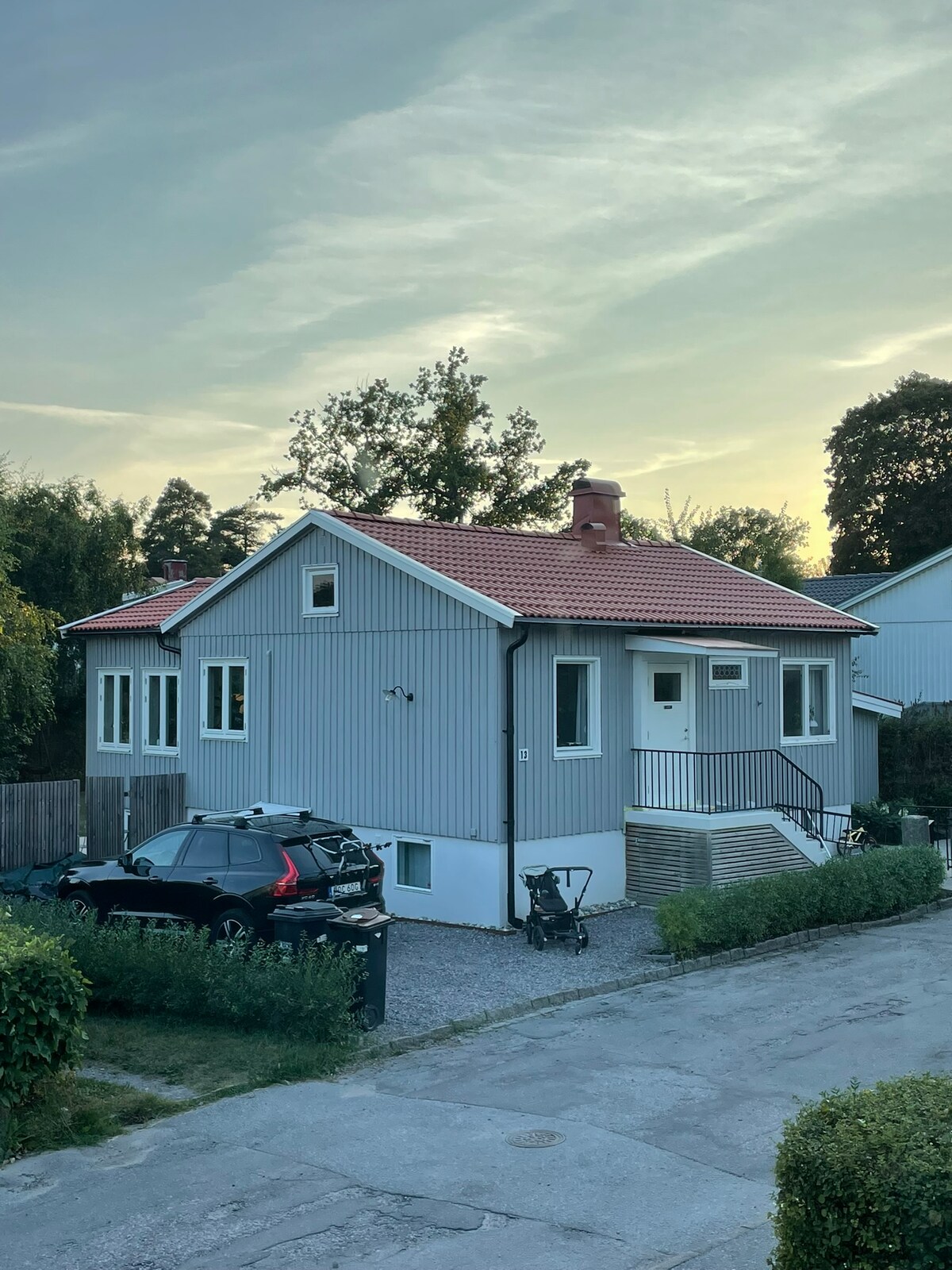 A charming gray house with a red-tiled roof is depicted, set against a serene sky at dusk. The property features multiple windows that provide natural light, and a gently sloping entryway leading to the front door. A driveway accommodates parked vehicles.