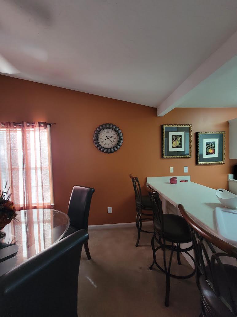 An inviting dining area features a round table with four chairs, complemented by a decorative centerpiece. A wall clock hangs above, with two framed art pieces in muted tones nearby. Warm orange walls provide a cozy backdrop, and a bar with stools is positioned against the kitchen.