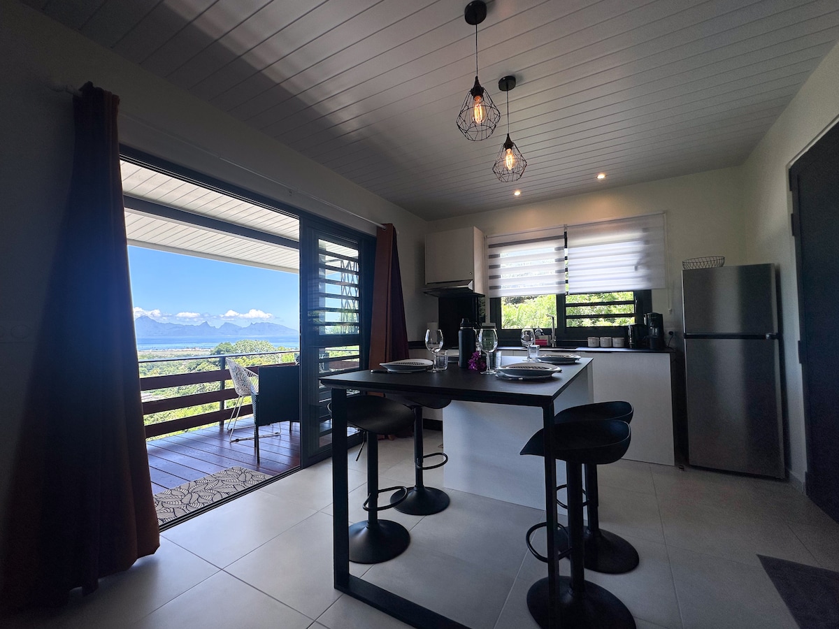 A modern kitchen area features a dining table with four black stools. Large glass doors open to a scenic terrace, offering a view of the mountains. Natural light is provided by overhead pendant lamps and daylight streaming through partially opened window treatments.