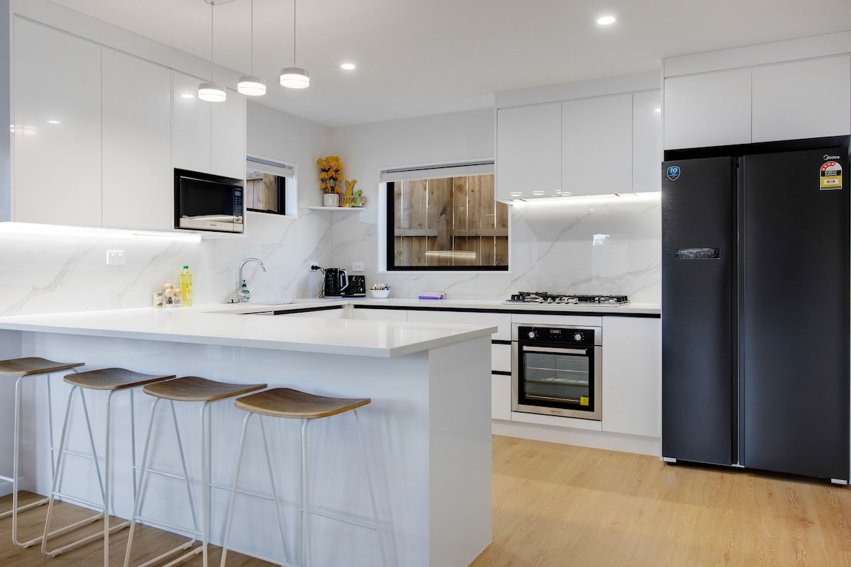 A modern kitchen features sleek white cabinetry, a large island with seating for four, and a stainless steel gas cooktop. An oven and black refrigerator are included, while a window provides natural light, enhancing the contemporary design.