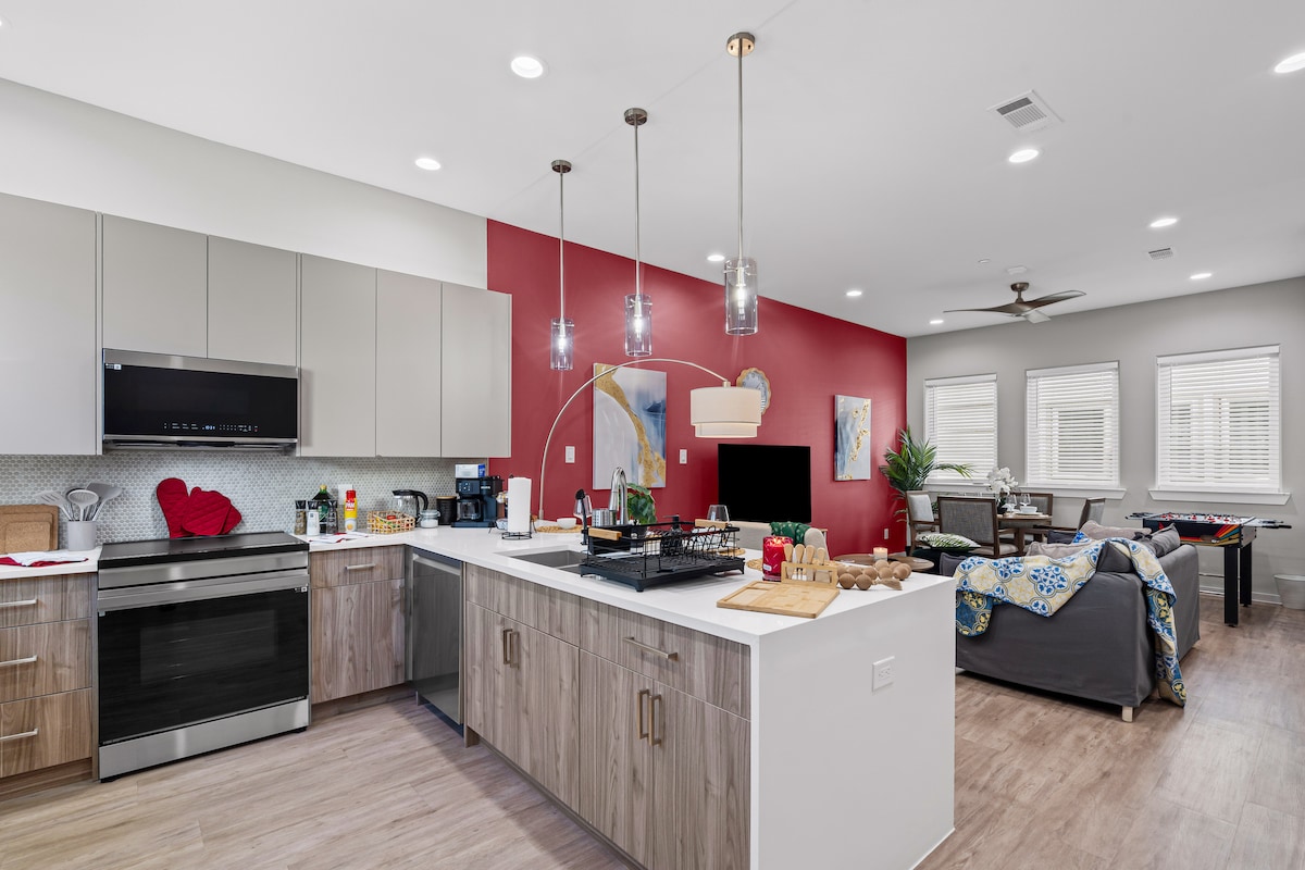 The kitchen features modern appliances, including a stove, microwave, and refrigerator, with sleek cabinets and ample countertop space. Pendant lights illuminate the area. A cozy living space is visible in the background, with a flat-screen TV and comfortable seating, creating an inviting open-concept layout.