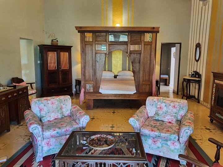 Discover a slice of old-world charm at our Luxury Villa in Goa. This spacious living area, complete with a unique carved wooden bed frame and vintage furnishings, offers a truly memorable and relaxing stay.