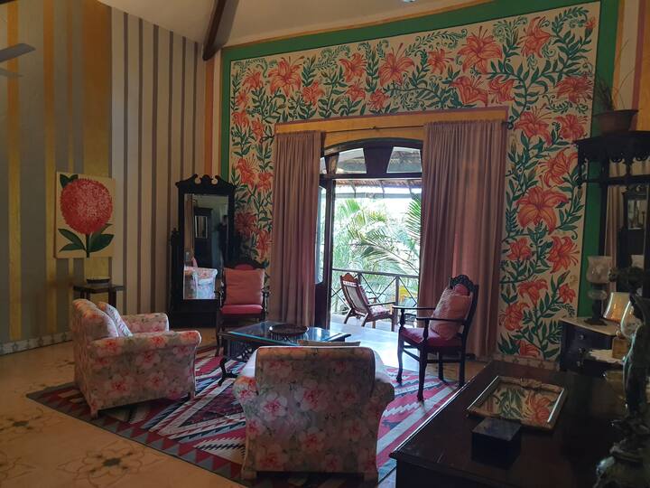 Step into a vibrant and elegant living room with a stunning floral mural, high ceilings, and a view of the lush greenery from the balcony. This is the perfect space to unwind and relax at our 6BHK Luxury Villa in Goa.