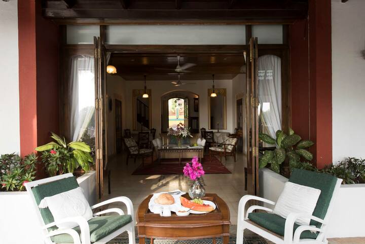 Discover the timeless elegance of our 6BHK Luxury Villa in Goa. This stunning living room, with its high ceilings and vintage wooden furniture, offers a beautiful space to relax in style.