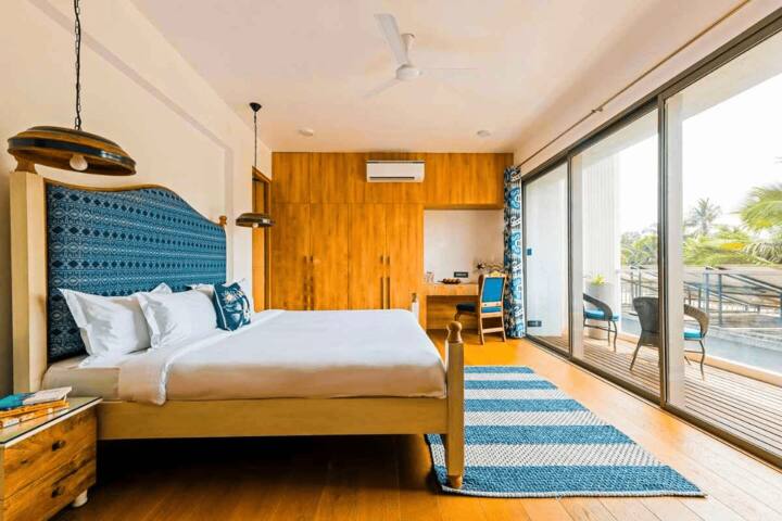 This spacious Goa villa bedroom offers a serene escape with a private balcony, stylish décor, a dedicated workspace, and ample natural light for a refreshing stay.