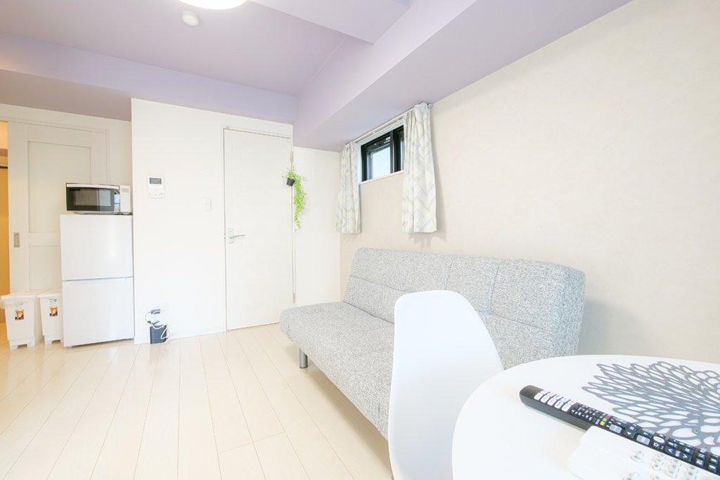 A well-lit living area is featured with a gray sofa bed, a small dining table, and a compact refrigerator. Light curtains frame the window, allowing in natural light. The space is designed with a neutral color palette, providing a calm atmosphere.