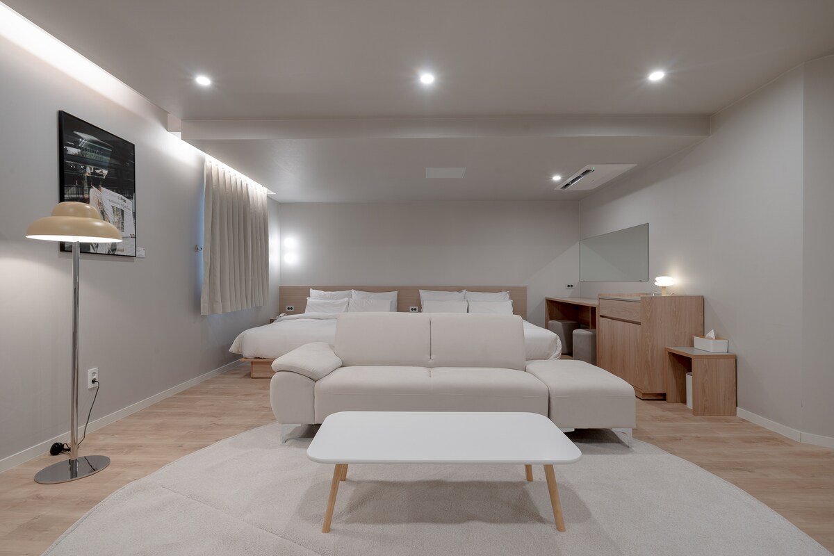 A contemporary living space features a light-colored sectional sofa and a low, white coffee table. Soft lighting provides illumination, while a large mirror and wooden desk are seen in the background. A subtle beige rug covers the floor, enhancing the warm ambiance.