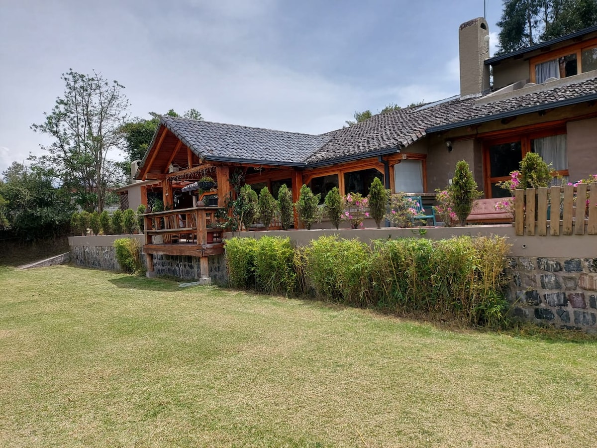 A large house features a wooden porch with a sloped roof, surrounded by neatly trimmed hedges and colorful flower beds. The spacious lawn extends in front, providing a serene outdoor area. Trees are visible in the background, contributing to the natural setting.