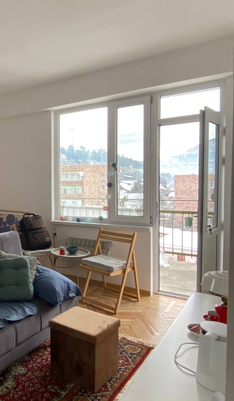 A bright living area is highlighted by large windows that open to a scenic balcony. A comfortable sofa is paired with a wooden coffee table, while a folding chair adds functionality. Soft textiles and a rug introduce warmth to the space, inviting relaxation.