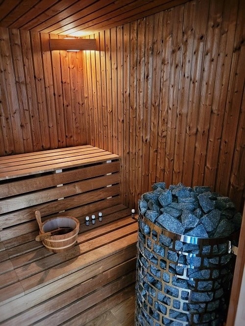 A wooden sauna interior features wooden benches arranged against the walls, complemented by a stone heater at the center. A bucket and small bottles are placed nearby, enhancing the rustic experience. Warm lighting illuminates the textured wooden paneling, contributing to a calming atmosphere.