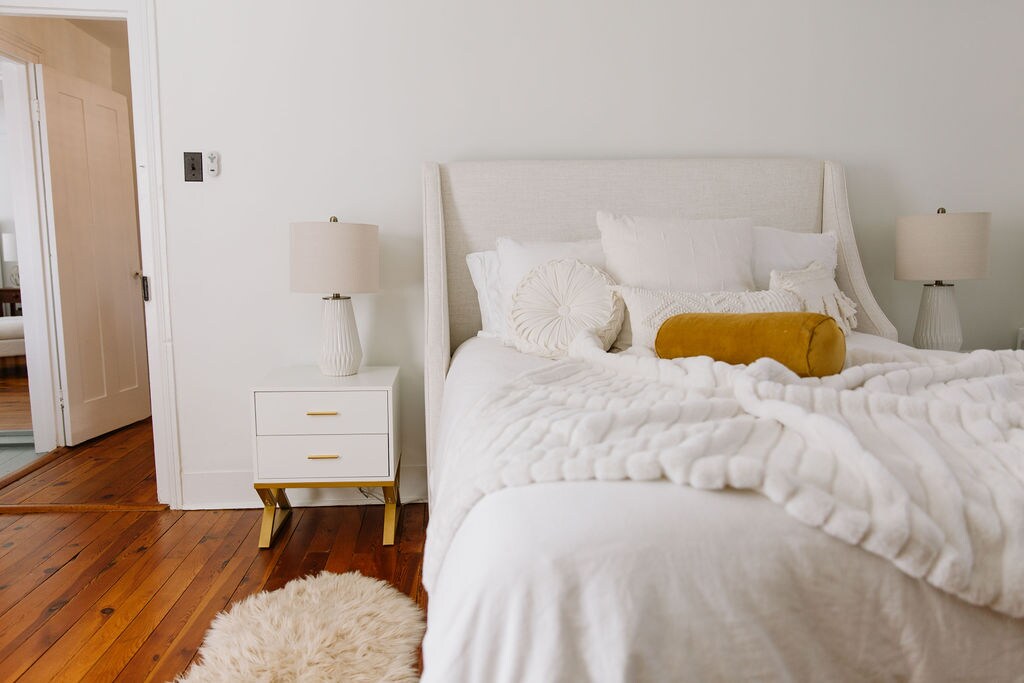 A spacious bed with layered white bedding and decorative pillows is positioned against a light-colored wall. A soft, round rug lies beneath the bed, accompanied by two bedside lamps on white nightstands. The entryway is visible in the background, featuring an open door.