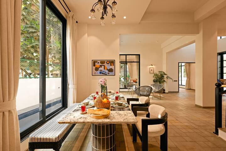 This elegant Goa villa features a spacious dining area with a stylish table and seating, bathed in natural light from large windows, perfect for memorable meals.