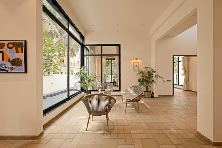 This Goa villa offers a bright, open space with floor-to-ceiling windows, inviting seating, and lush greenery, seamlessly blending indoor comfort with outdoor views.