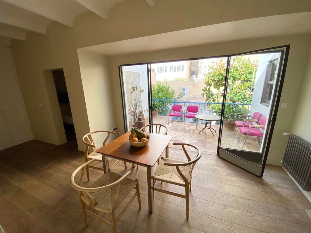 A dining area features a wooden table surrounded by four woven chairs. Large glass doors lead to a patio, showcasing pink chairs and a small table amidst greenery. Natural light brightens the room, enhancing the inviting space.