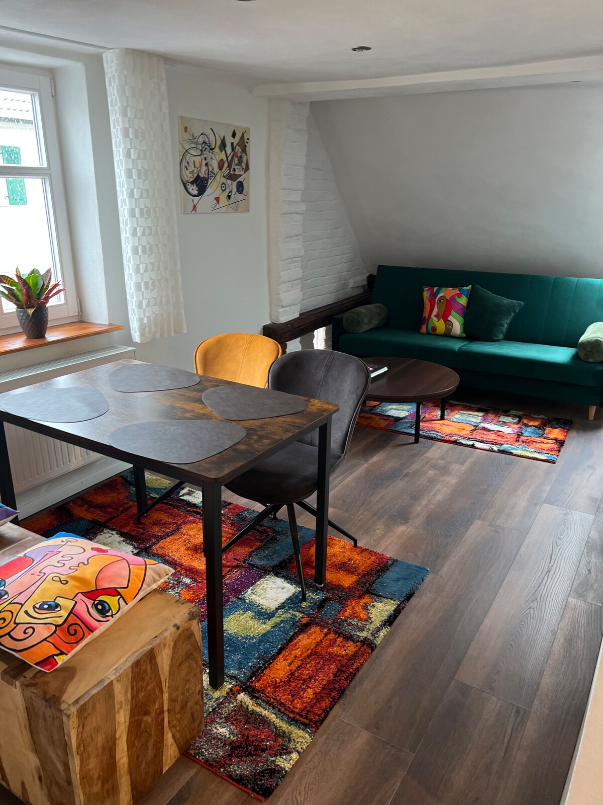 A bright and inviting living area features a small dining table surrounded by chairs, complemented by a cozy couch. Colorful rugs enhance the space, while artwork adorns the walls, creating a visually stimulating environment. Natural light floods in through a window, adding warmth to the room.