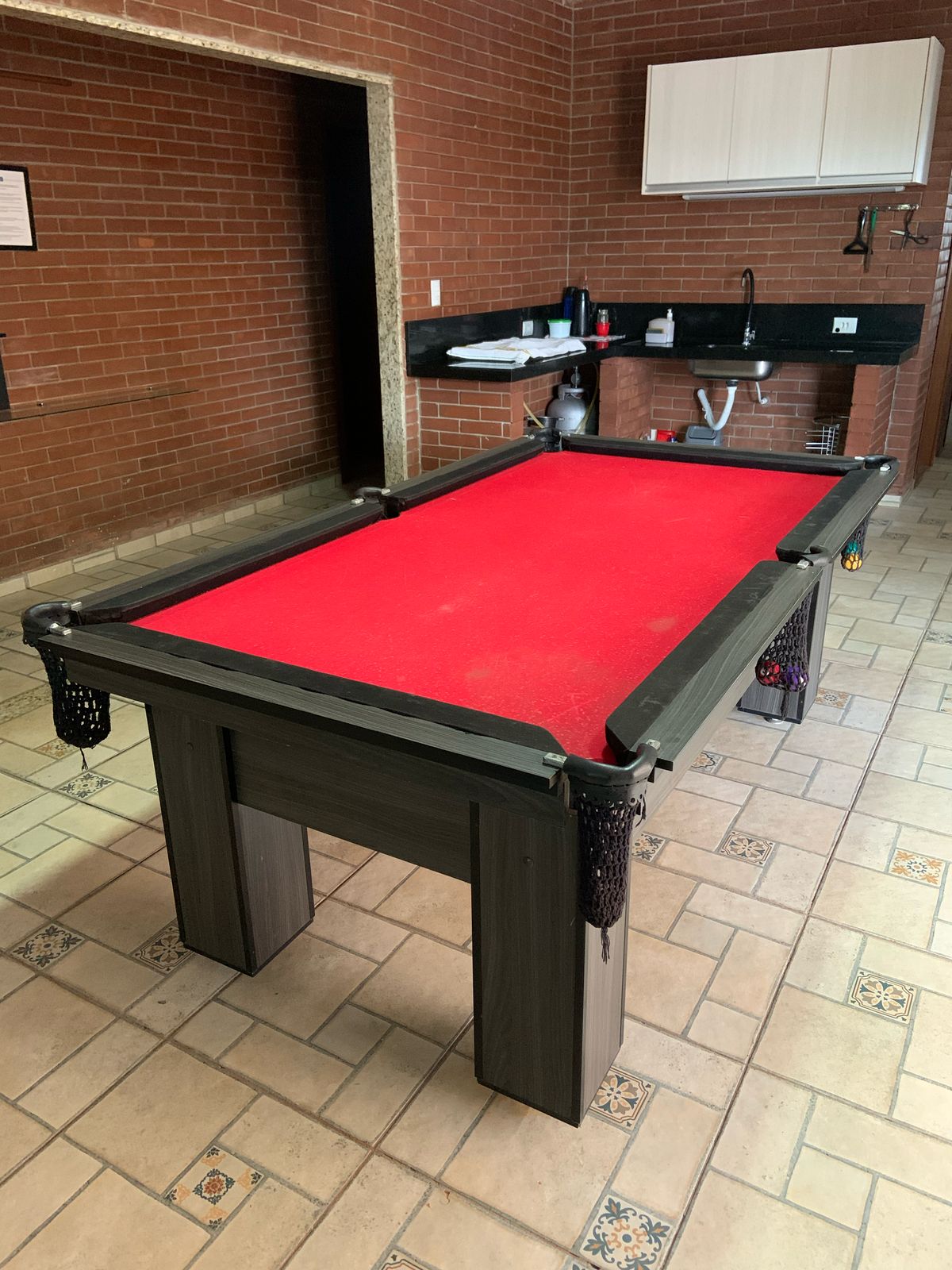 A billiard table with a vibrant red felt surface is positioned in a spacious area. The table features a sturdy frame with pockets at each corner. A modern kitchen area with black countertops is visible in the background, complemented by brick walls and tiled flooring.