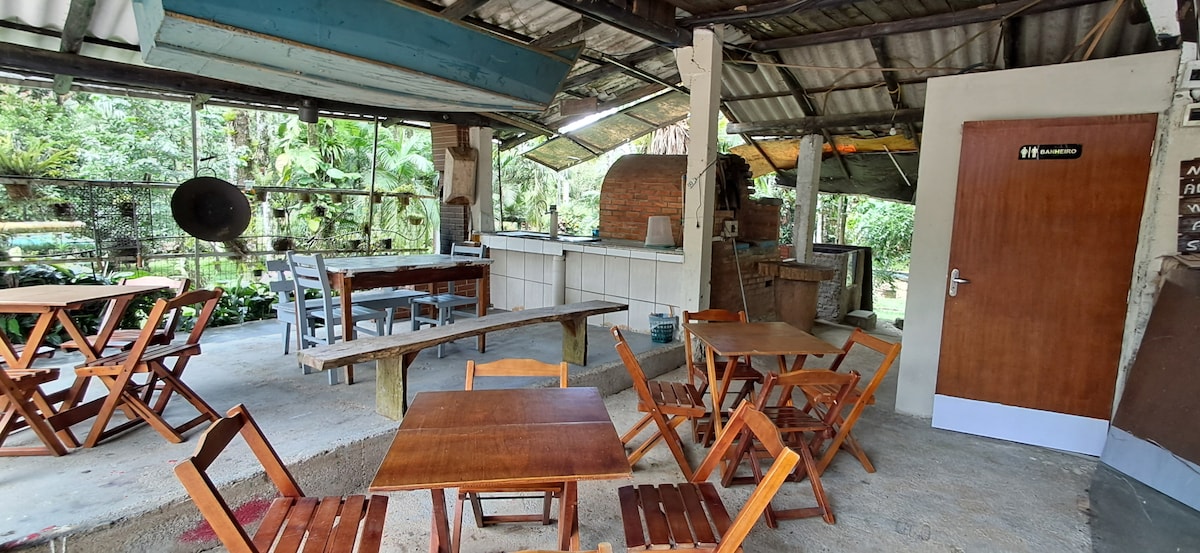 An outdoor dining area features wooden tables and chairs arranged around a central table. A rustic kitchen area with a brick oven and an open workspace is visible. Large windows allow natural light to illuminate the space, creating a connection to the surrounding greenery.