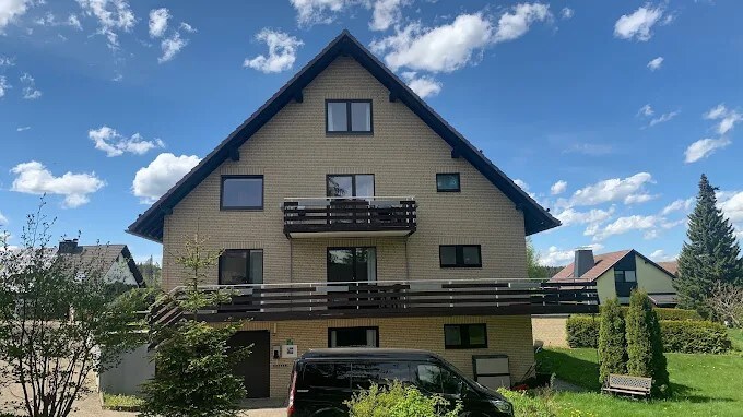 A multi-story building is displayed with a triangular roof and balconies. Large windows provide ample natural light. A black vehicle is parked in front, and greenery is present with shrubs and small trees flanking the entrance. The sky is bright with scattered clouds.