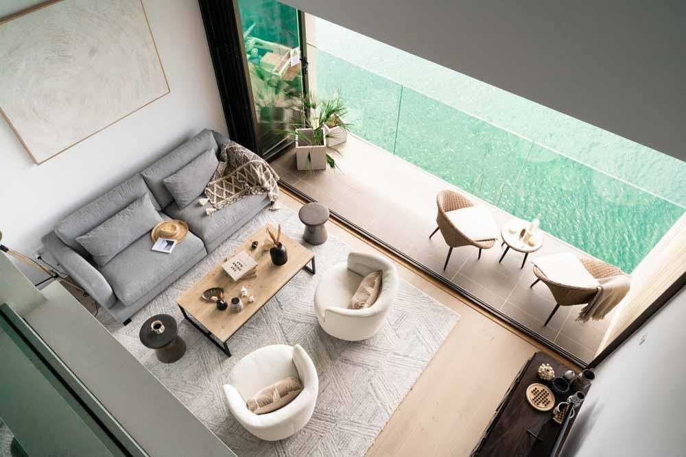 An open-plan living area features a cozy gray sofa and two white armchairs positioned around a central coffee table. Floor-to-ceiling glass doors lead to a private balcony with outdoor seating, showcasing views of the calm estuary waters beyond.