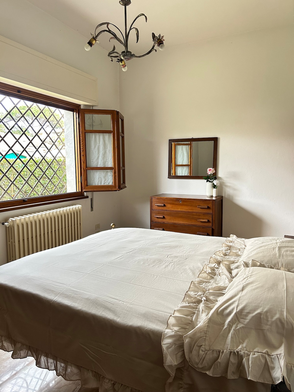 A serene bedroom features a spacious bed adorned with a soft, ruffled bedspread. A wooden dresser is positioned against a wall, accompanied by a small mirror reflecting soft natural light. Two windows with open shutters provide a view of the outdoors, enhancing the room's brightness.